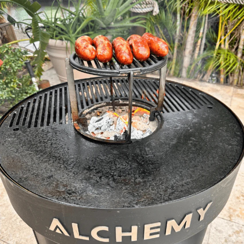Alchemy Plancha grill with sausages cooking on a metal stand, surrounded by greenery.