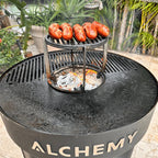 Alchemy Plancha grill with sausages cooking on a metal stand, surrounded by greenery.