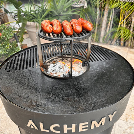 Alchemy Plancha grill with sausages cooking on a metal stand, surrounded by greenery.